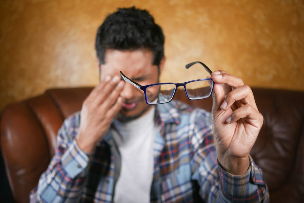 A man in plaid shirt holding eyeglasses indoors. Focus on eyewear with neutral background.